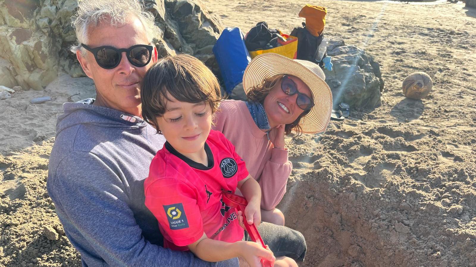 Image of a white haired man, wearing sunglassses, digging a hole on a beach with a young boy sitting on his lap holding a beach spade and a woman with a sunhat on and sunglasses sitting in the background.