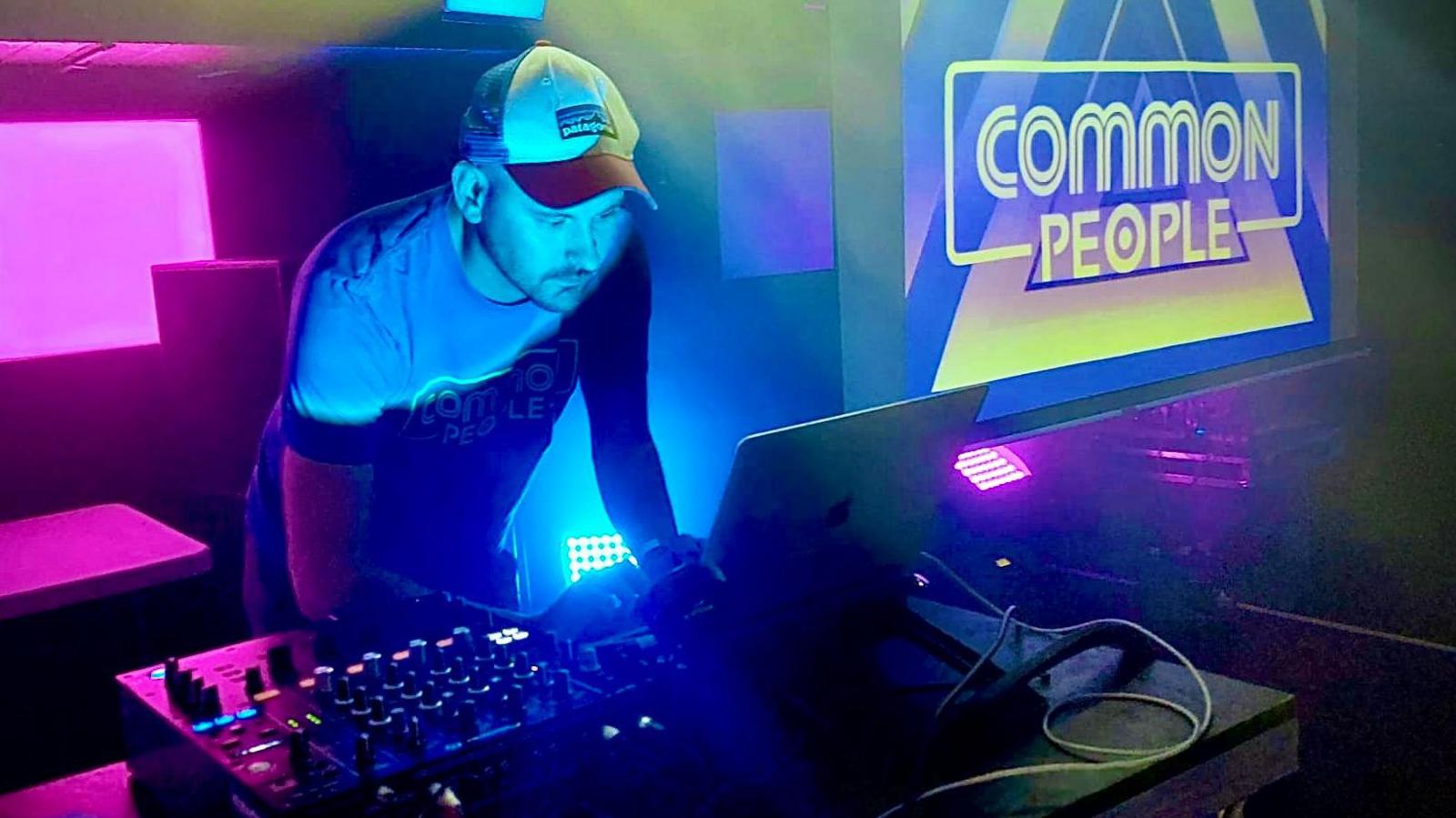 Jon Rogers is wearing a baseball cap and a Common People-branded T-shirt while leaning over a DJ console. The desk is lit up in neon lights and an illuminated Common People sign is to the right.