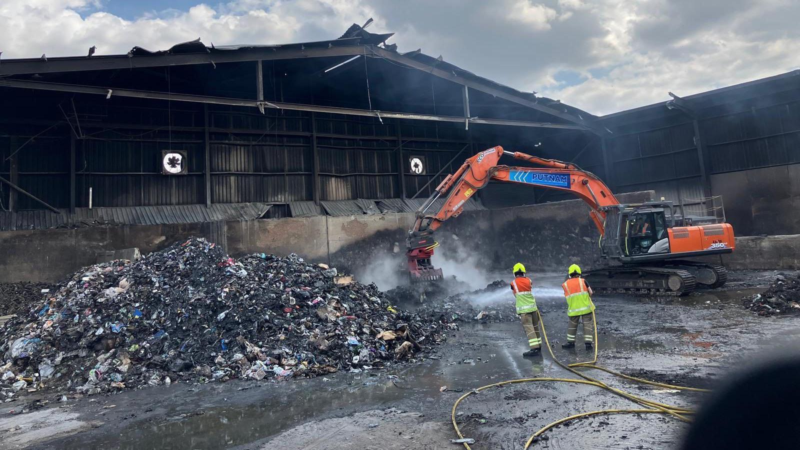 A burnt out waste site, with a crane, to the right, smoke coming from the rubbish, two fire crews holding water hoses, piles of rubbish to the left and a burnt out building behind.