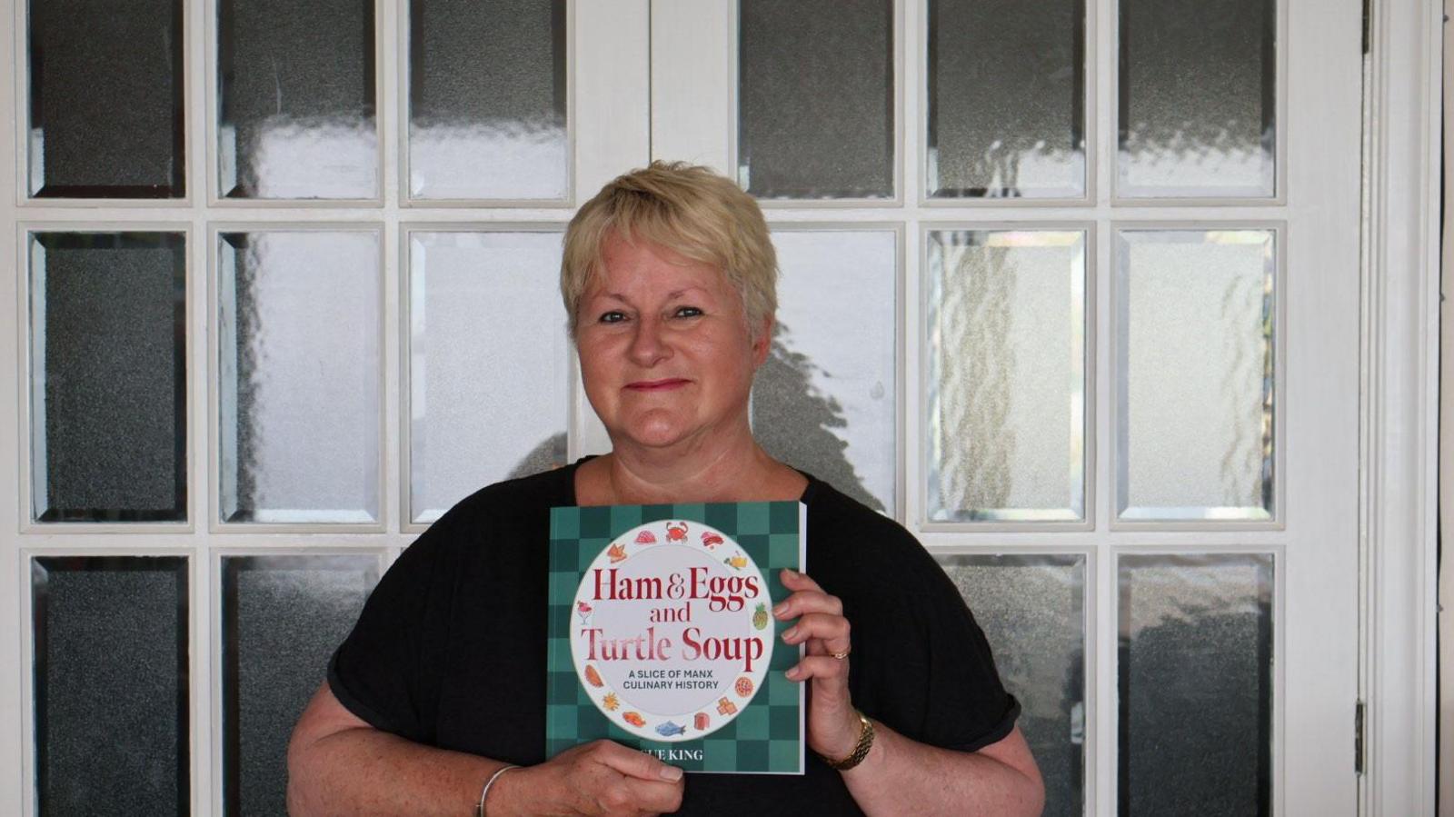Sue King, who has short blonde hair, is photographed wearing a black t-shirt. Standing in front of panelled window doors, she is smiling while holding up a copy of her book. Primarily green, the cover reads Ham & Eggs and Turtle Soup.