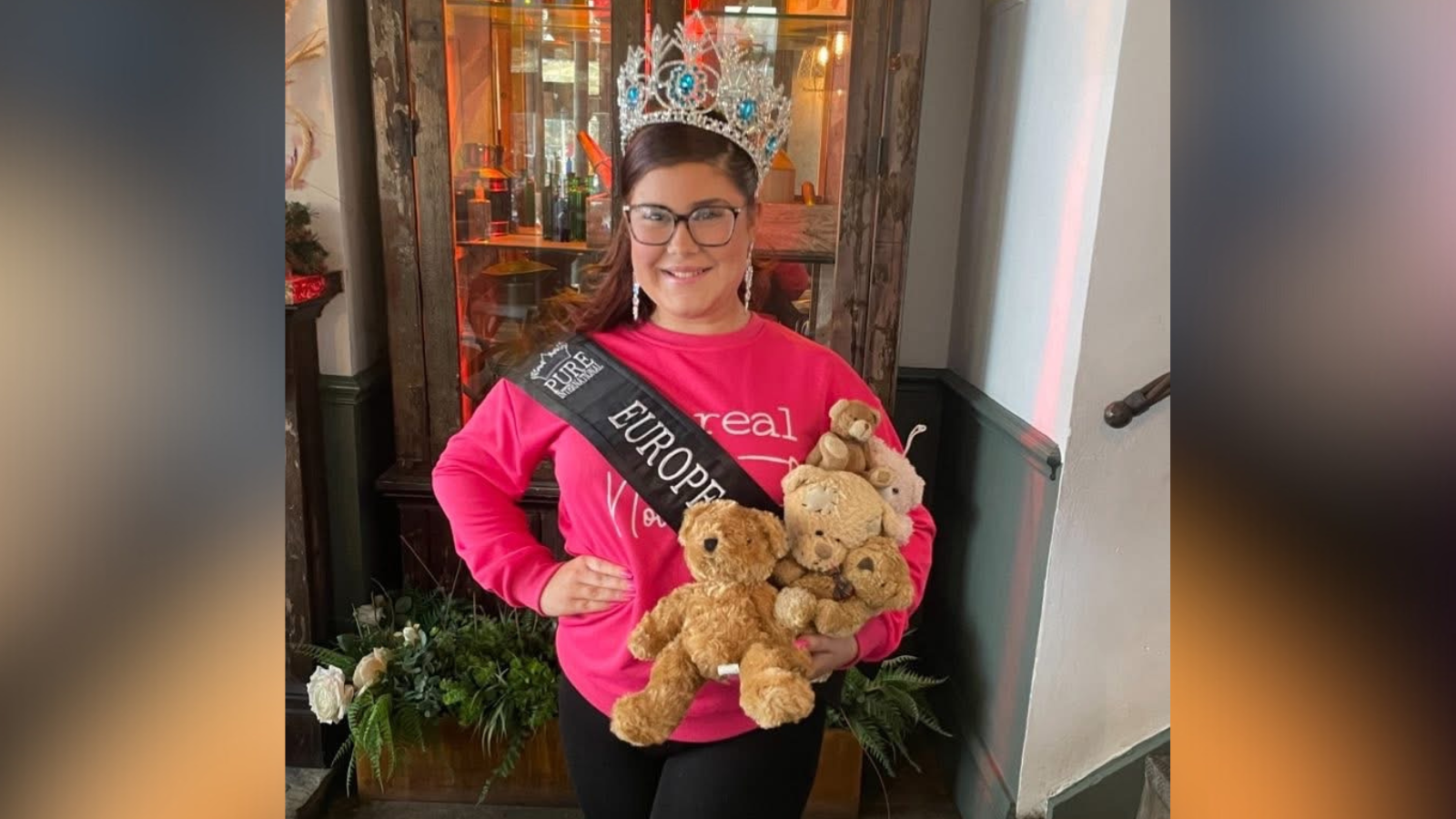 Bella is stood wearing her crown and a sash and big silver earrings. She is holding a handful of teddies in one hand and her other hand on her hip.