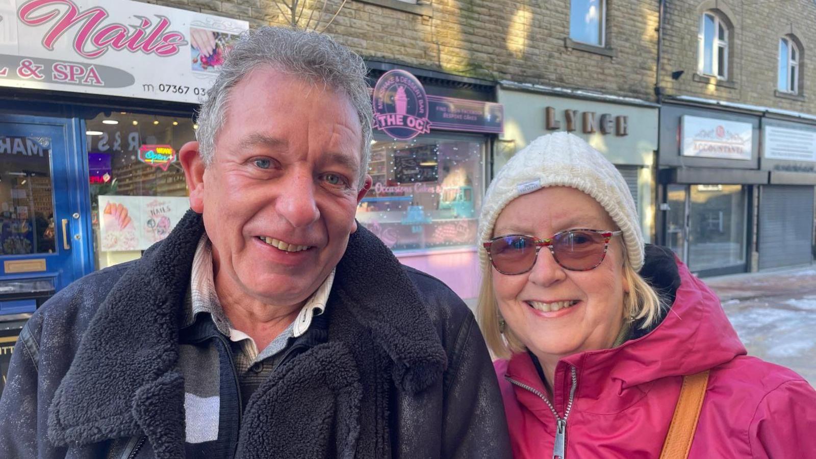 A man wearing a blue fleece jack and woman wearing a pink coat, glasses and a hat are standing in front of a row of shops in Elland.