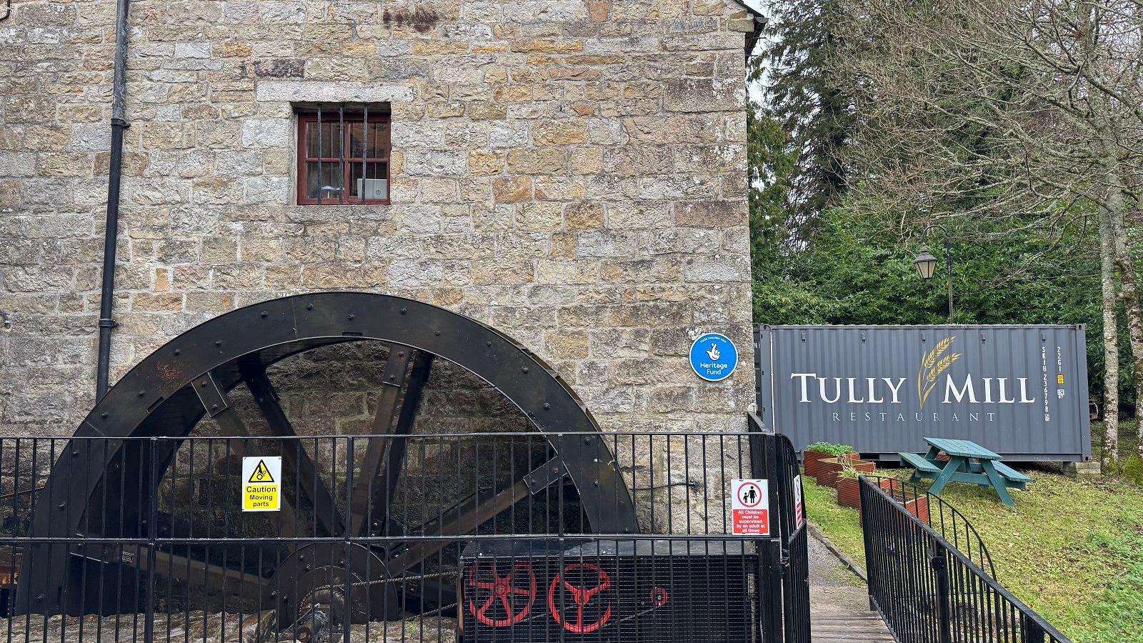 The waterwheel of an old mill. The words Tully Mill Restaurant are visible on a shipping container in the nearby yard.