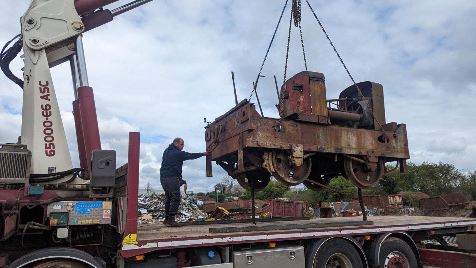 An old rusty locomotive is lifted onto a truck bed. It is being lowered on to it while a worked guides it down. 