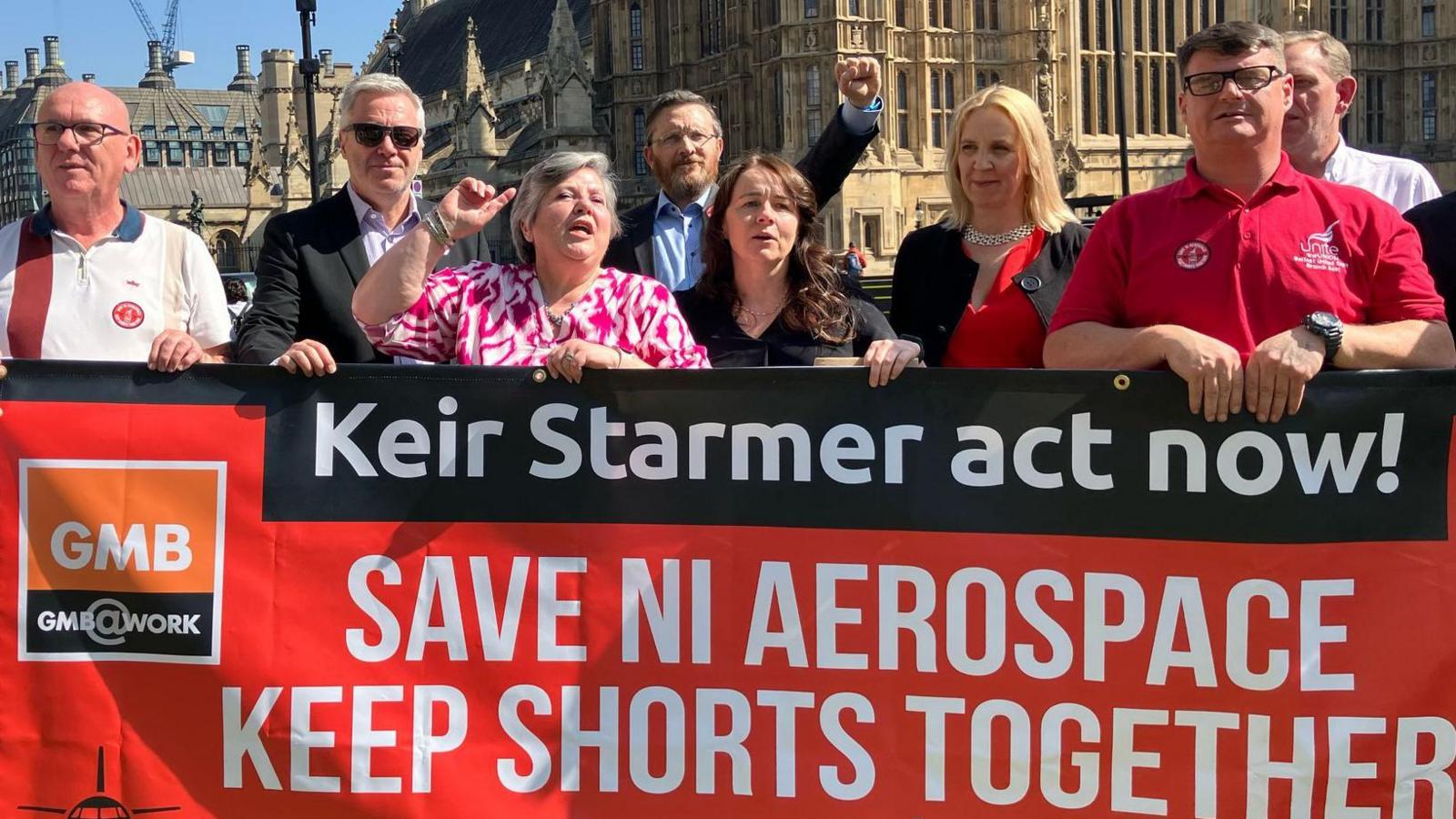 A group of protesters, stood outside Westminster, holding red GMB union banners that say "Keir Starmer act now! Save NI Aerospace keep shorts together... We're stronger together, save NI aerospace"