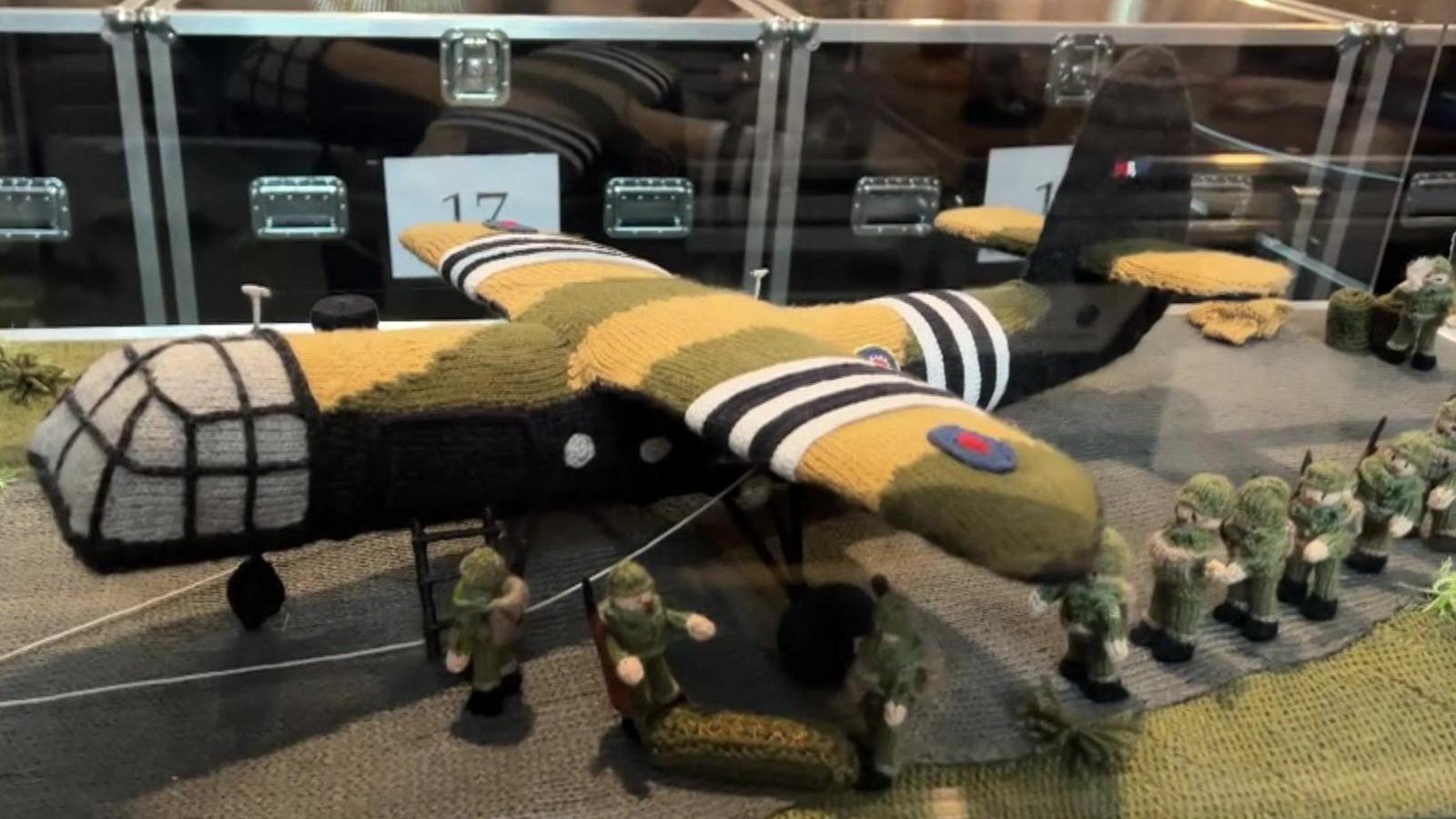 A knitted green plane, with knitted soldiers lining up next to it.