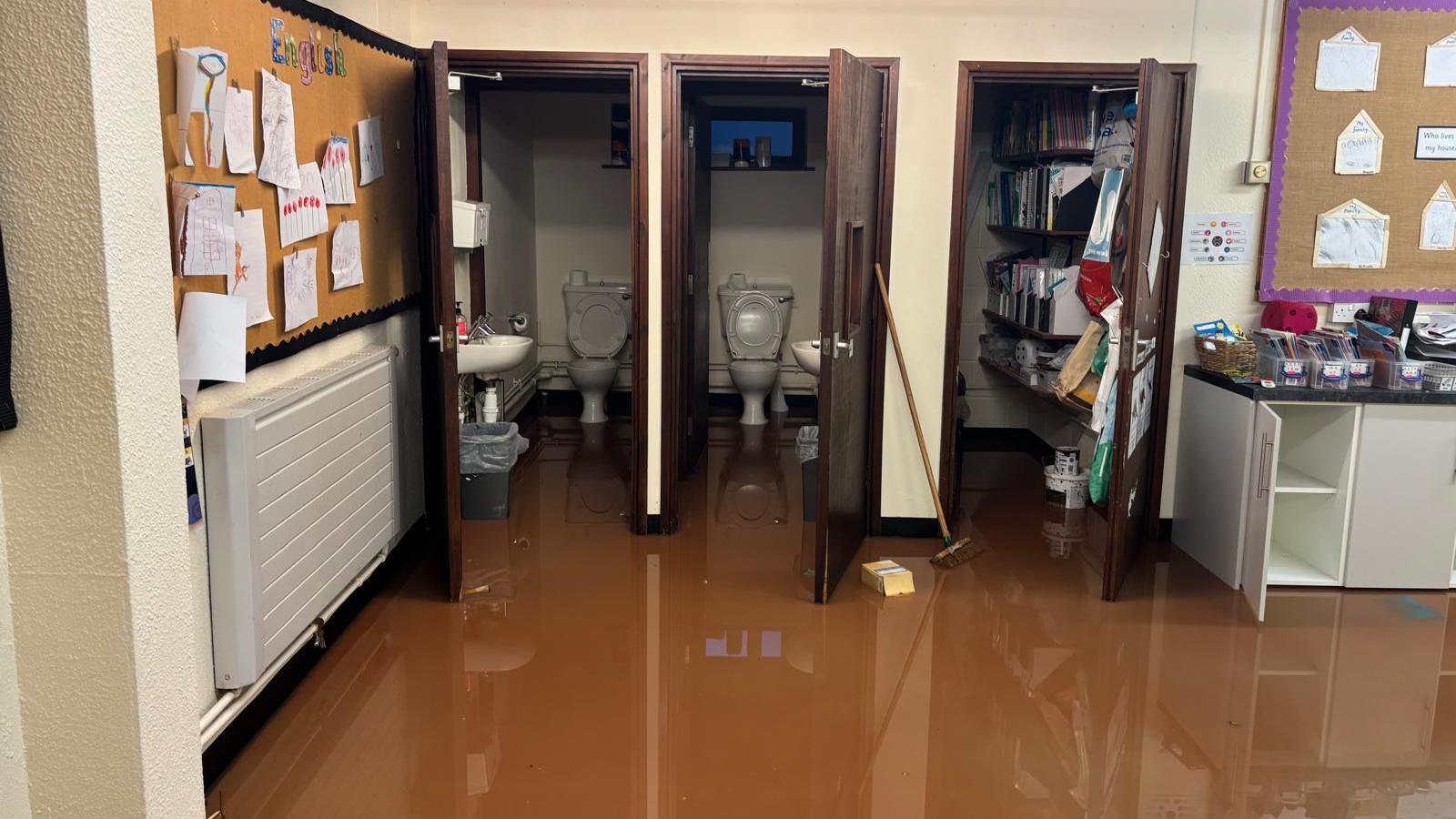 A classroom with two toilet cubicles and a cupboard filled with books and folders has brown water throughout it.