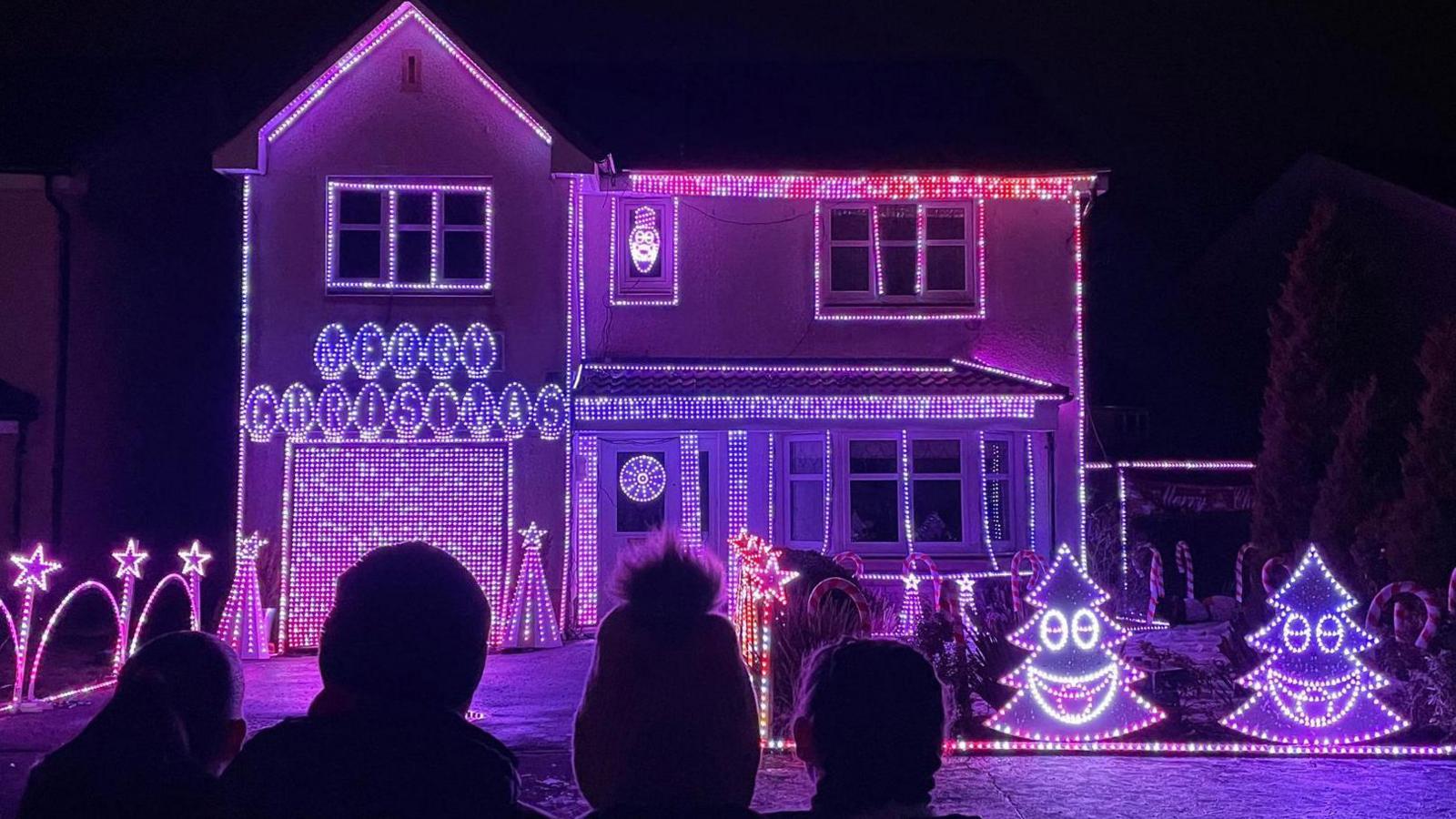 A two-storey home illuminated in bright pink and purple Christmas lights. There are small LED Christmas trees with smiley faces and the driveway is bordered by brightly lit archways and stars.