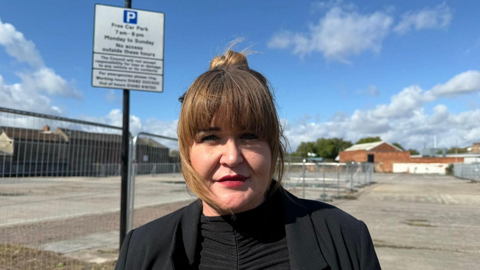Kerry Ryan smiling at the camera. She has brown hair which is tied up into a bun on the top of her head. She also has a fringe, and is wearing a black blazer and black top. She's standing in front of an empty car park, which has lots of metal railings. There is a sign above her head with parking information on.