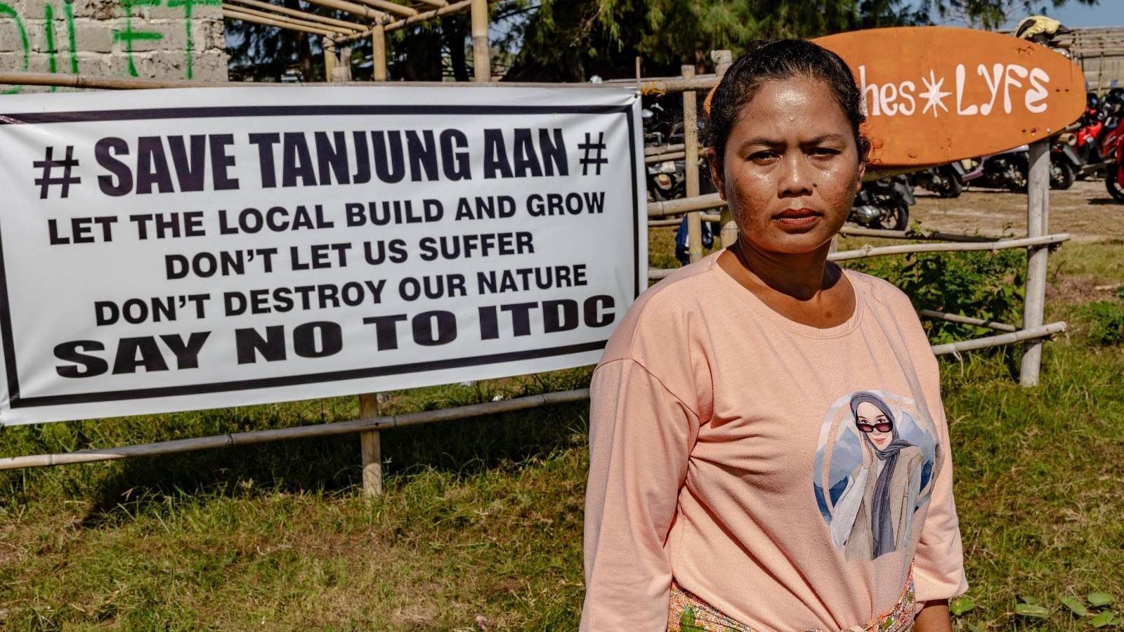 Ella Nurlaila in a peach coloured long-sleeved shirt, looking into the camera with a burrowed eyebrows. Behind her is a large banner that reads: "Save Tanjung Aan, let the local build the grow, don't let us suffer, don't destroy our nature, say no to ITDC".