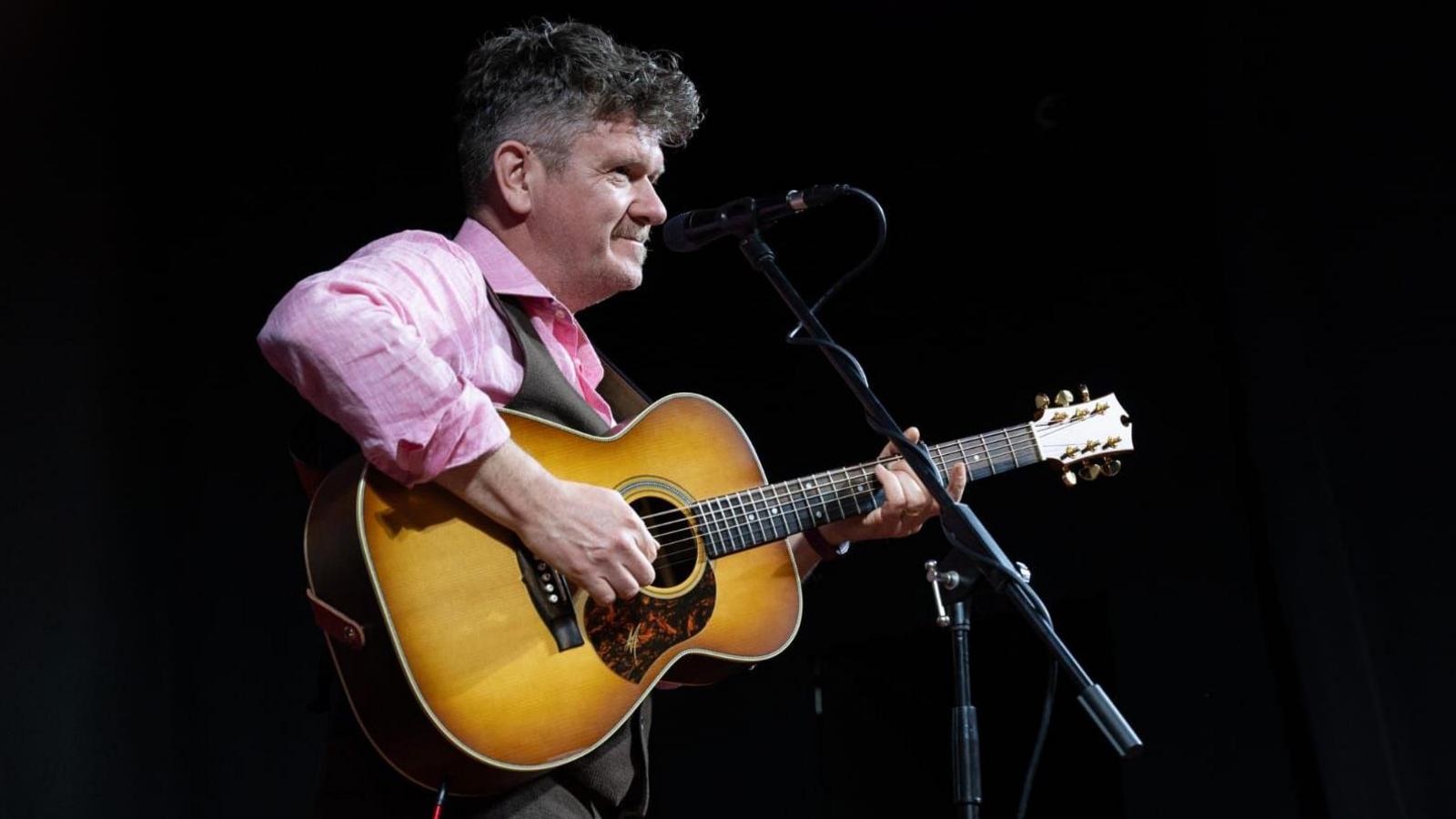 a man with a guitar at a microphone. He is lit as though he is on stage and he is wearing a pink shirt