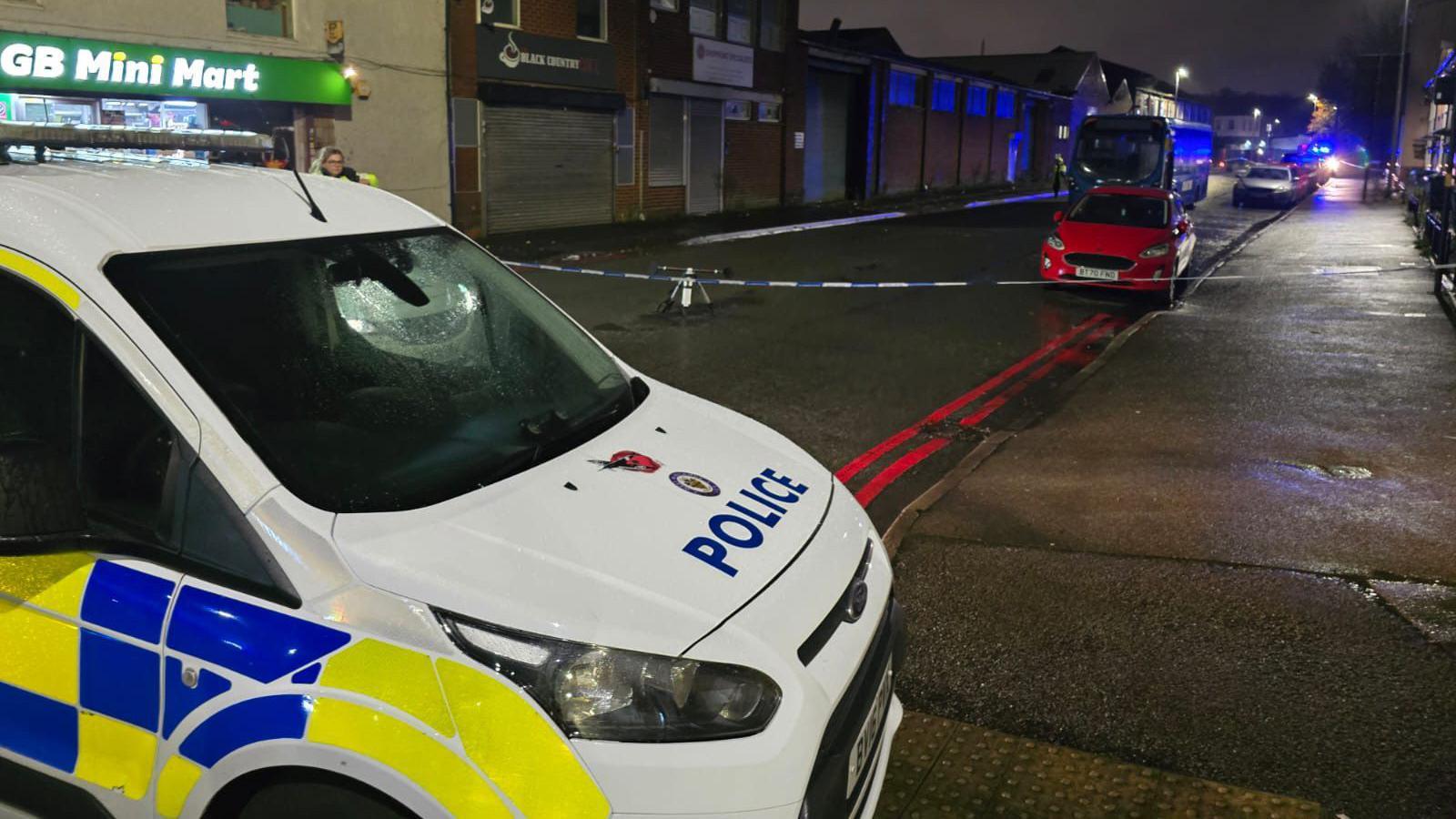 A street with a police cordon. The sky is dark and street lights are on. There is a police car at the front of the image, with a police cordon, a red car, and a blue bus behind it on the street. There is a corner shop with a green and yellow sign on the left of the street.