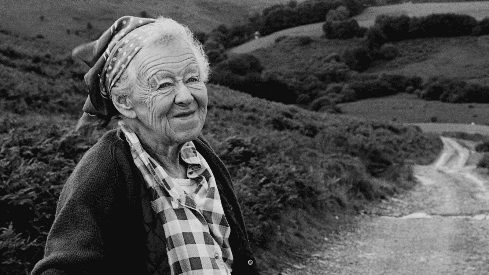 A black and white photograph of an elderly lady with white hair wearing a head scarf, a checked blouse and cardigan, with a road through Exmoor behind her