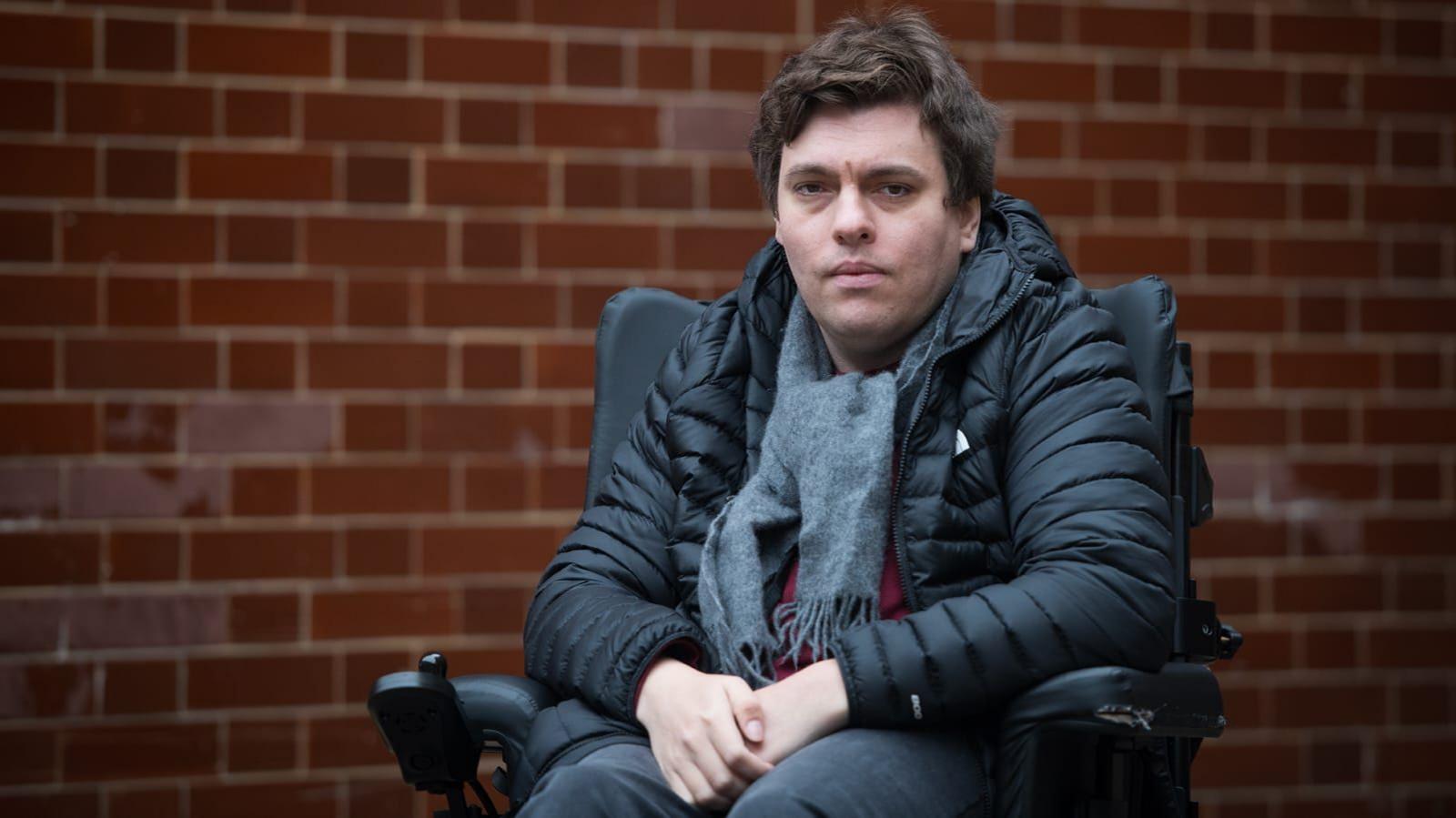 Alex Taylor, a white man wearing a black jacket and grey scarf and in a wheelchair, on a London street