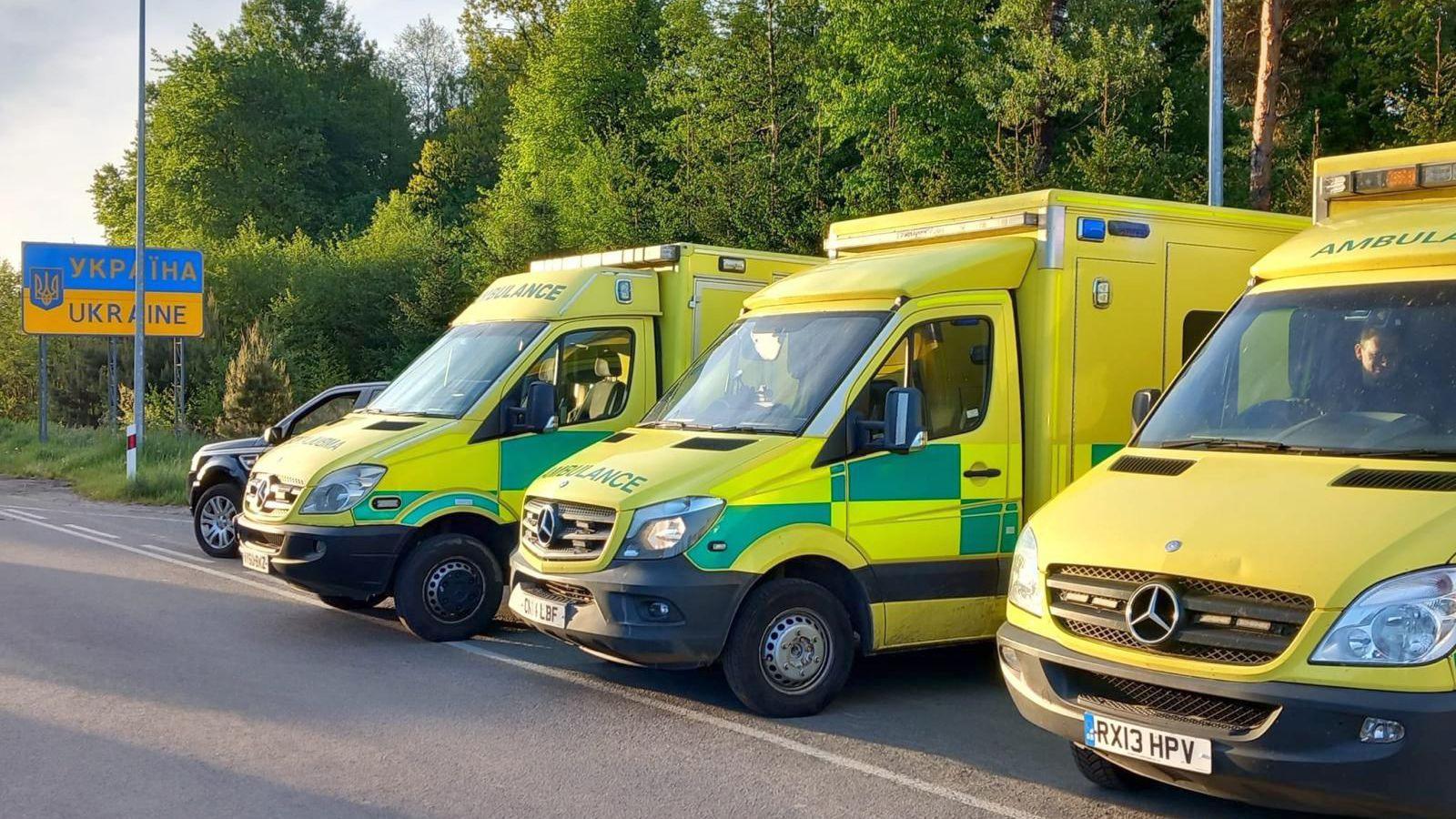 Three lime green ambulances lined up at the crossing point between Poland and Ukraine. The rectangular Ukraine sign blue and orange halves.