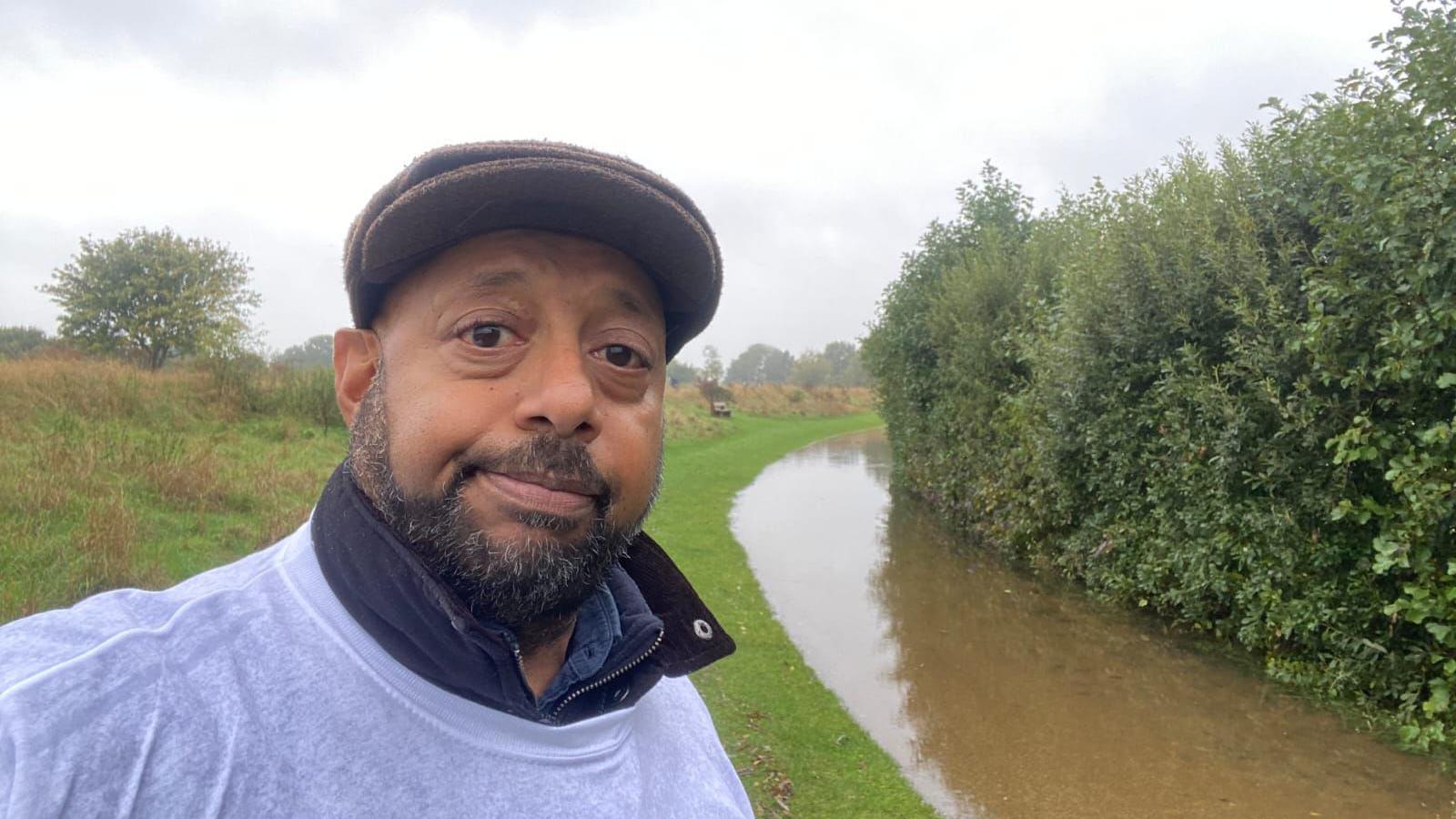 Asif Shahid's from Sue Ryder Peterborough's team takes a selfie wearing a white T-shirt and a cap. He has a sad expression and behind him is a flooded footpath. 