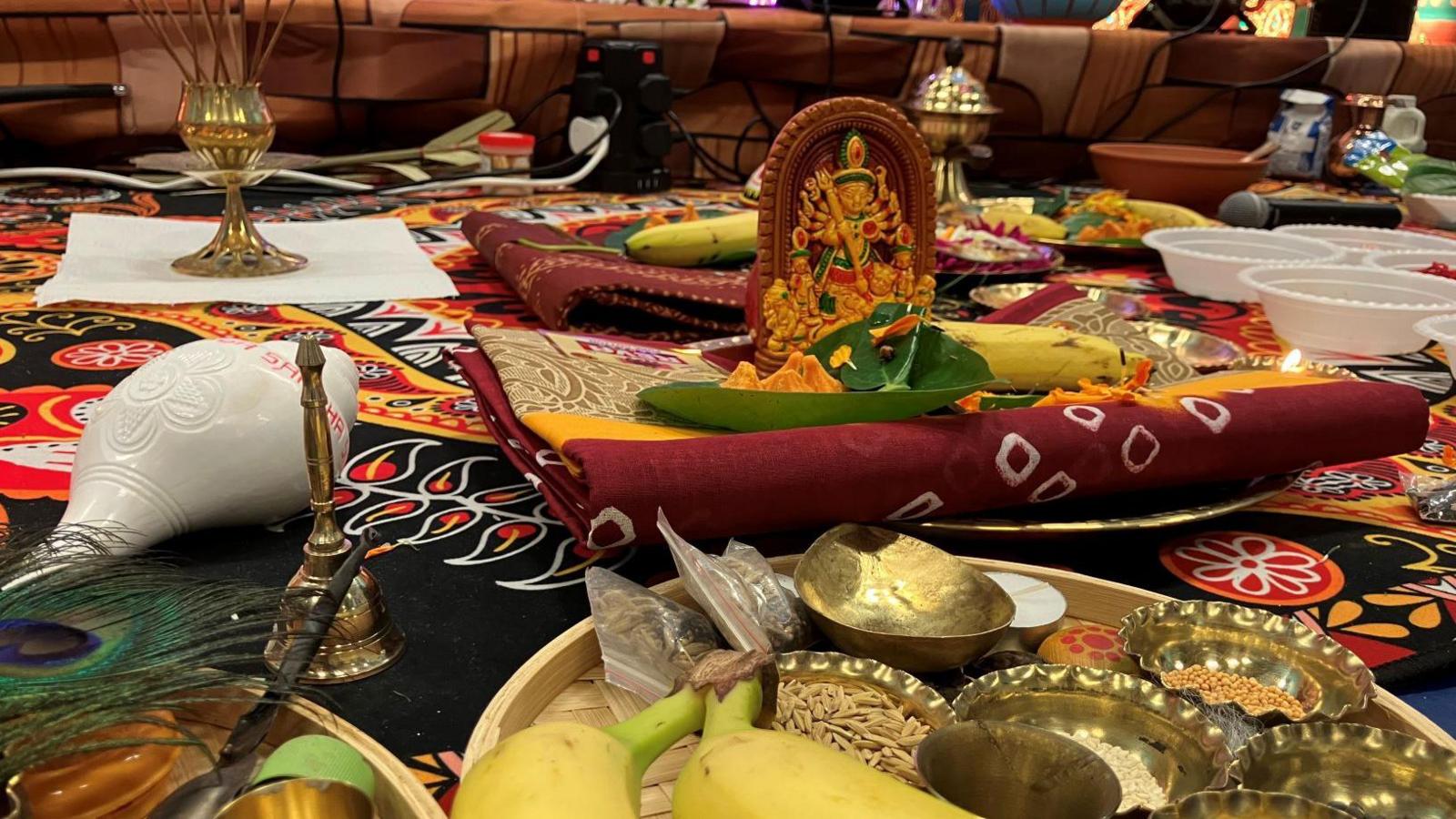 a small icon of the Hindu goddess Durga on some folded material, with bananas and bowls of seeds on a tray. There is a white conch shell and incense sticks in the background.