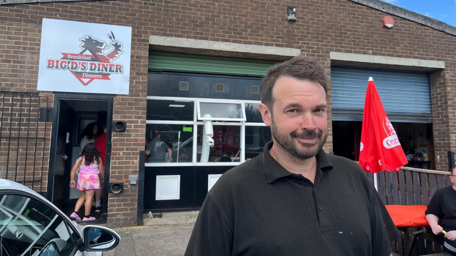 A man with brown hair and a beard, wearing a black polo shirt, stands in front of the cafe under a sign that reads: 'Big D's Diner'.