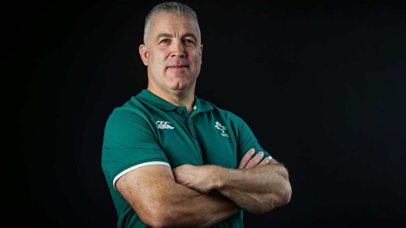 A man with short cropped light hair stands facing the camera with his arms crossed. He is wearing a short-sleeved green Irish rugby top