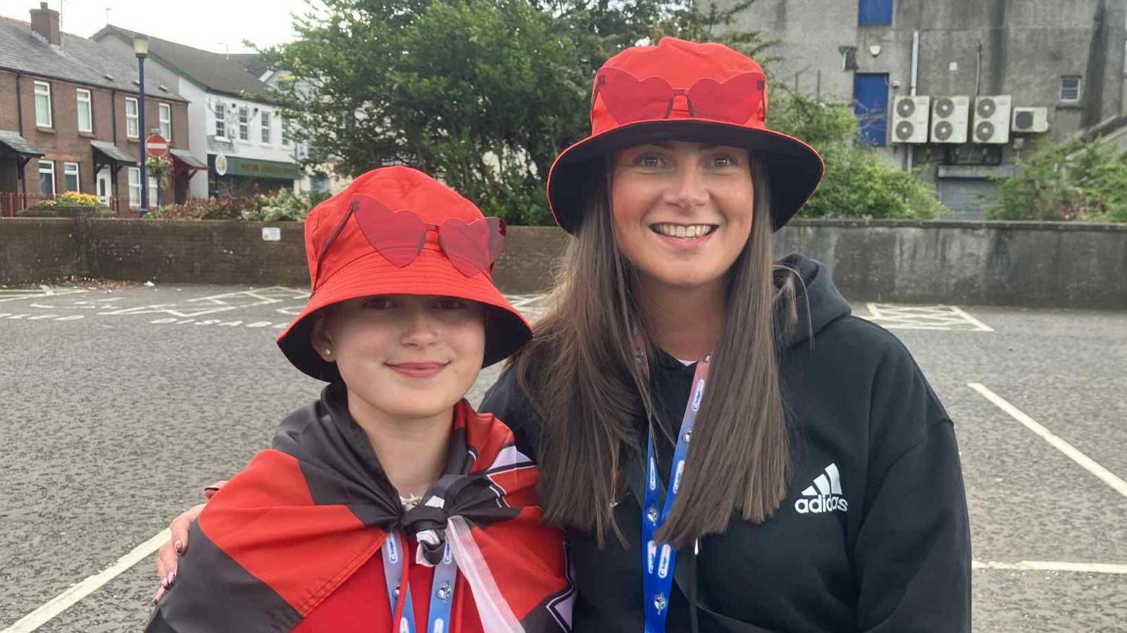 A woman and a small girl stand side by side and are smiling. They are wearing red hats and have heart-shaped glasses resting on them. The young girl is wearing a red and black crusaders flag tied around her shoulders.