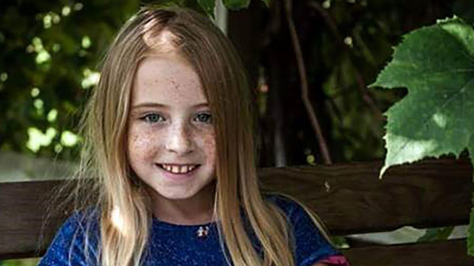 Cimarron Thomas is a young girl with long strawberry blonde hair and lots of freckles on her face. She is smiling in the photo, sitting on a bench with lots of green leaves around her. 