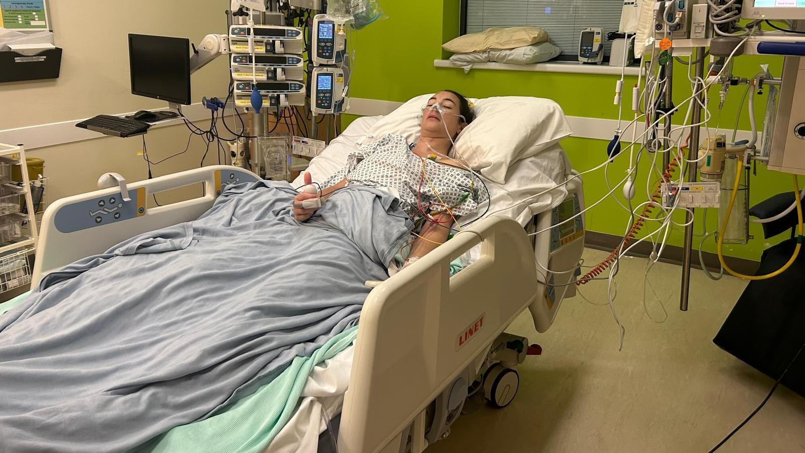 Bex Hind is lying on a hospital bed wearing a hospital gown. She has many tubes attached to her, which stretch to various machines. She looks unwell but is giving a thumbs up to the camera.