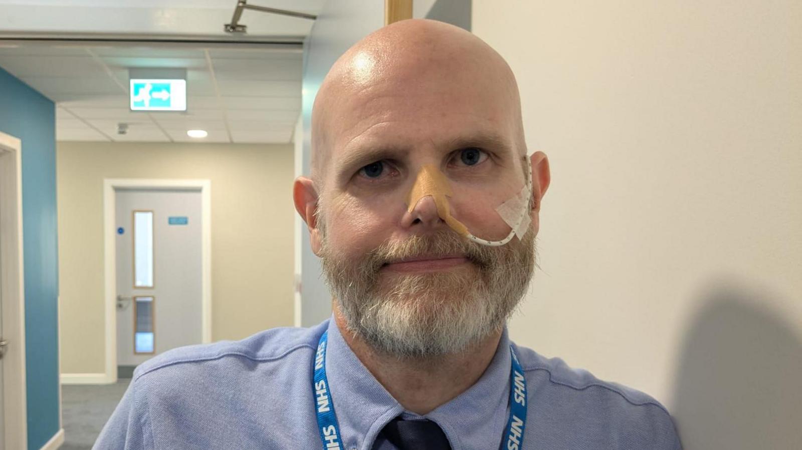 Gareth, a bald, white man with a thick greying beard leaning on a white wall. He is looking into the camera with a neutral look on his face. He is wearing a light blue shirt with a navy blue tie, and a blue NHS lanyard. He has a tube that goes into his left nostril with plasters holding it in place. The background includes a hallway with white and blue walls, white doors and a neon green fire escape sign.