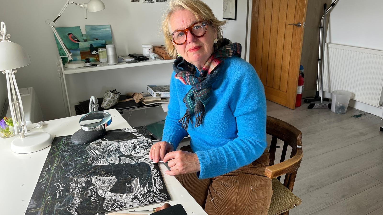 A woman wearing a blue jumper and a woollen scarf sits in an artist's studio. She is at a desk working on a black-and-white picture of puffins. 