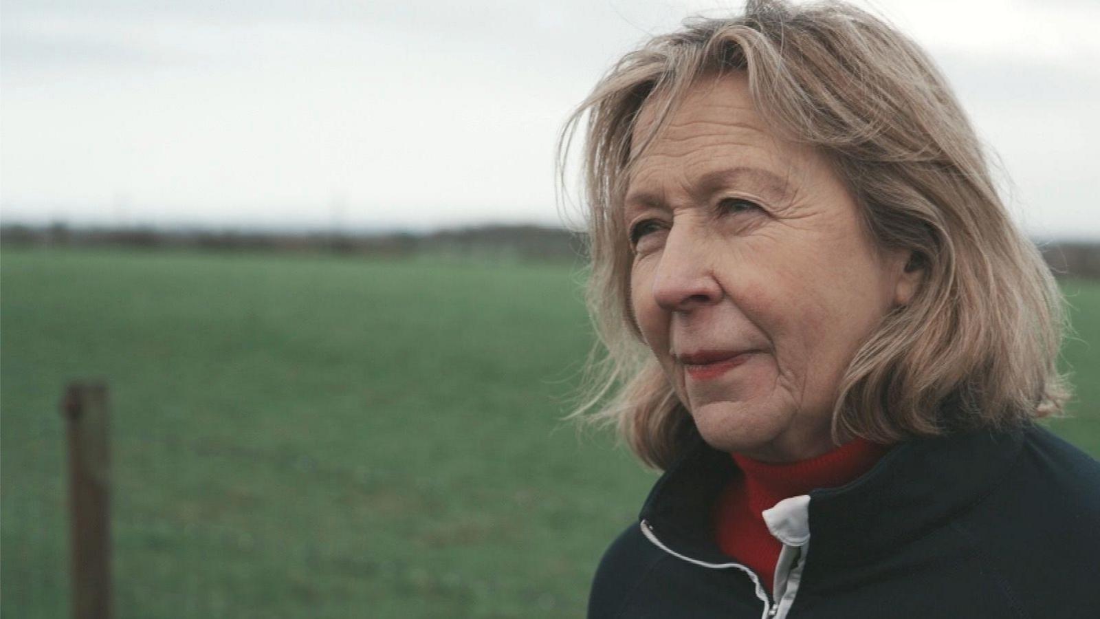 Chris Pollitt, with shoulder-length blonde hair, wears a red polo-necked jumper and a black cardigan as she stands in front of a green field.