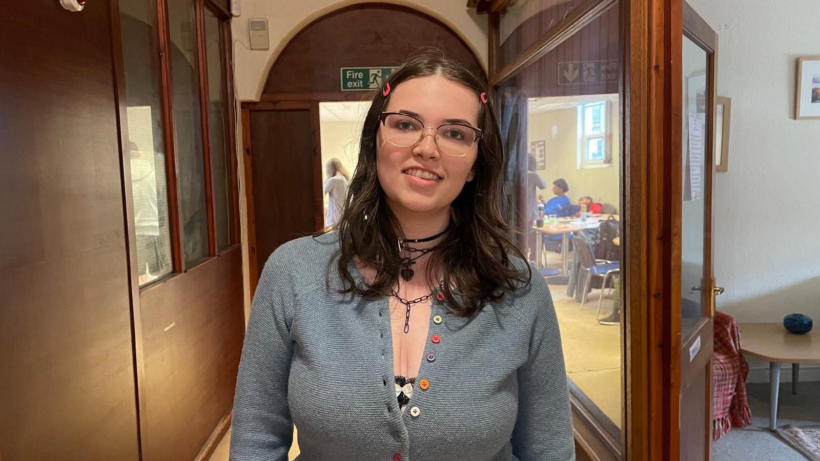 A woman in a grey cardigan with dark, long hair. She has glasses and two pink clips in her hair. She is stood in a corridor with a room behind her. 