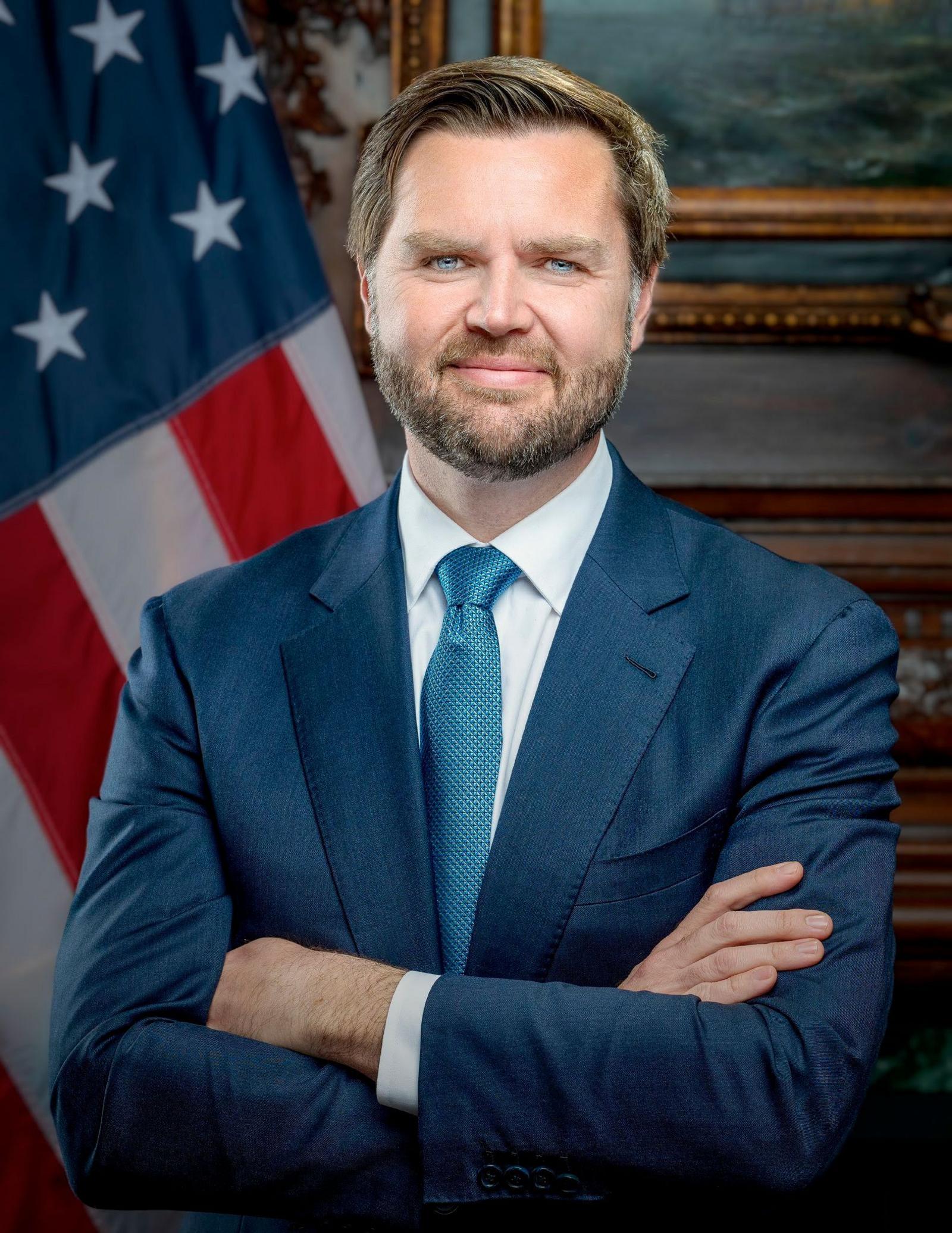 JD Vance stands with his arms crossed and a slight smile, wearing a blue suit and tie.