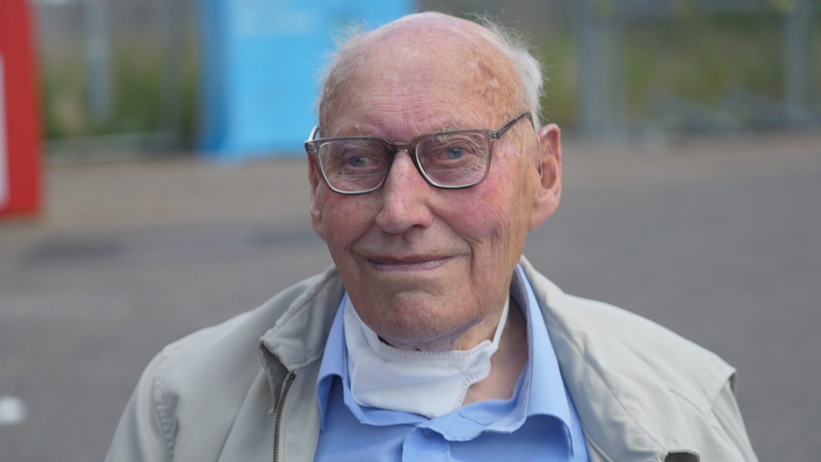 A man with a blue shirt and glasses. He is smiling at the camera and wearing a cream coat 