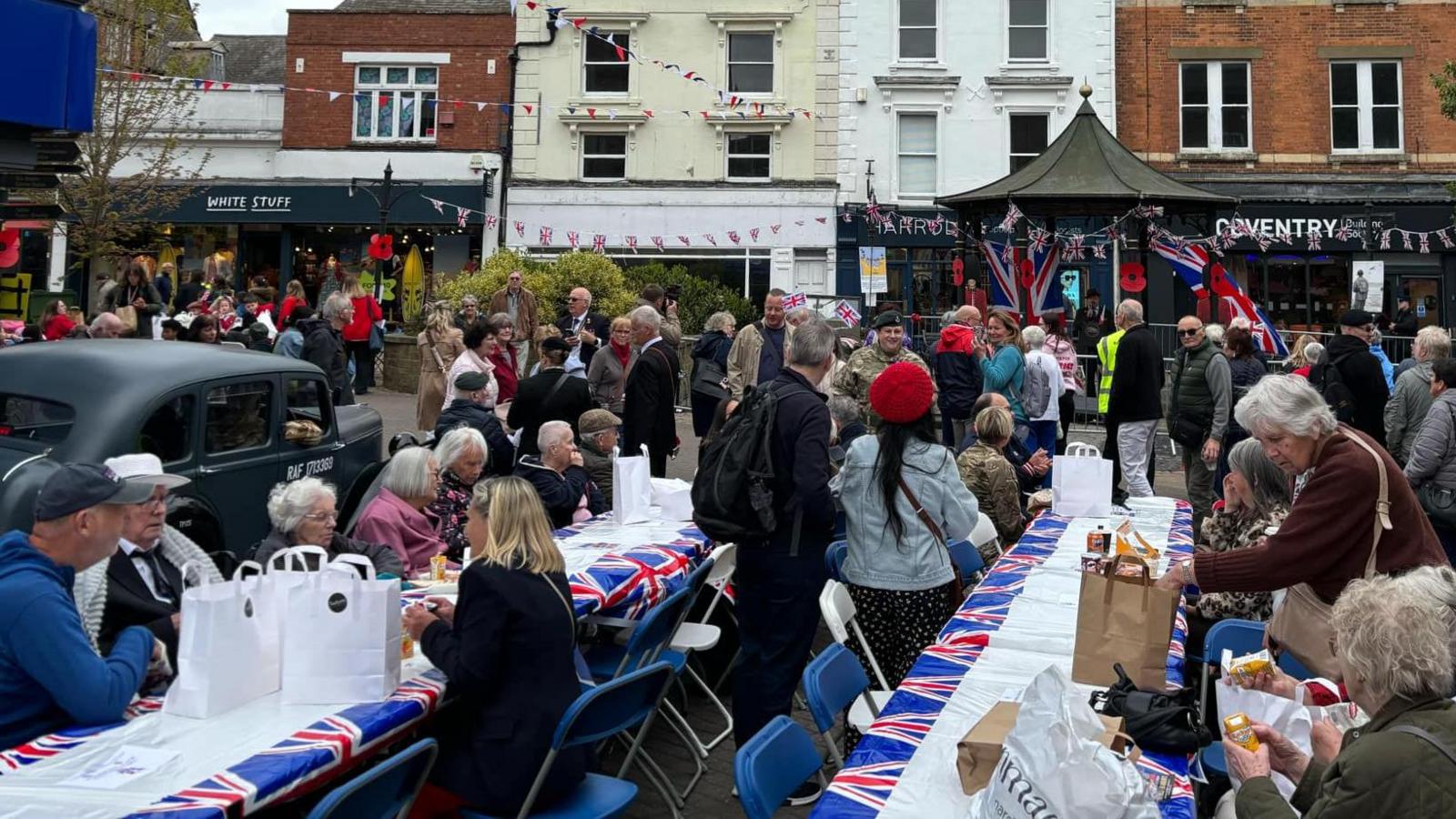 Dozens of people sitting at long tables on the pedestrian area outside town centre shops. The tables are decorated with union flag table cloths.
