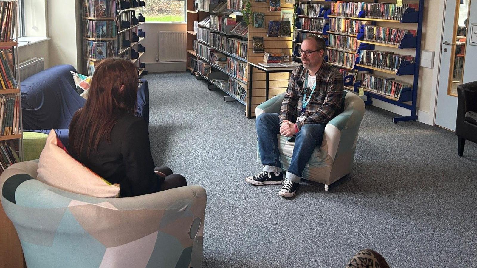 A man sitting across from a girl in a chair. In the background is rows of bookshelves.