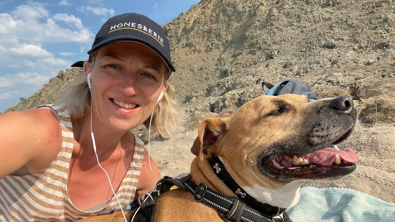 Bayliss is smiling in next to her dog at Holywell Bay. Bayliss is wearing a black cap, headphones and a striped vest.