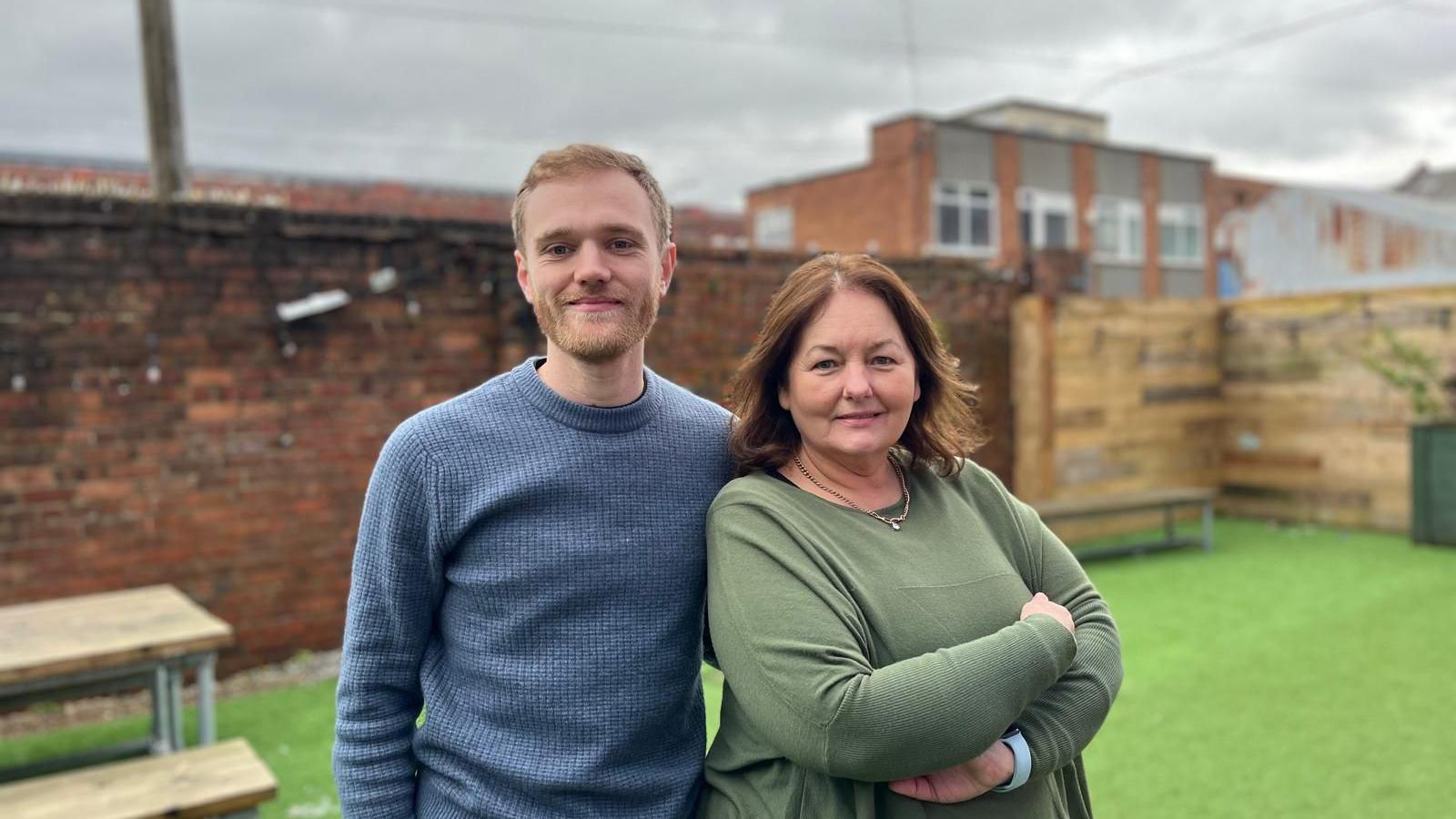 Joseph Burns, with sandy coloured hair and a light beard and wearing a blue sweatshirt, and Julie Maddocks with brown hair and a green top, smile at the camera in a beer garden area with tables and benches in the background.