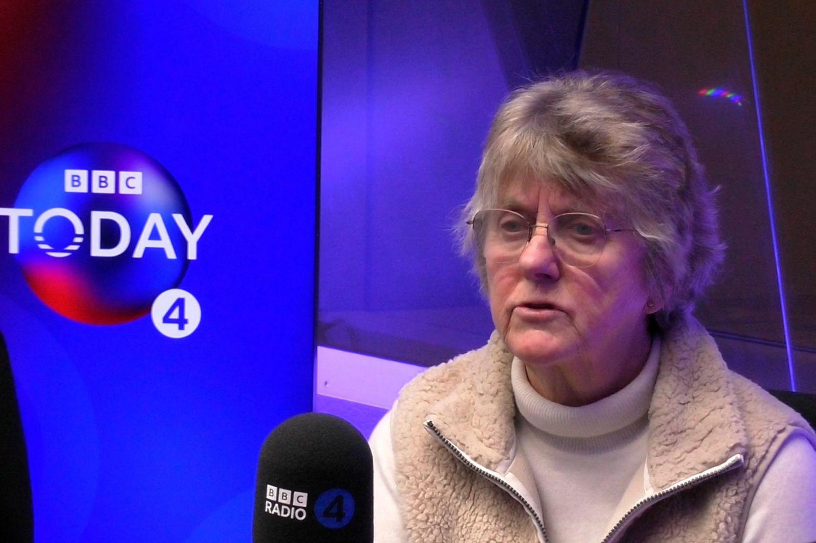 A woman with short grey hair and glasses is sat in a radio studio. She is speaking into a microphone labelled "BBC Radio 4"