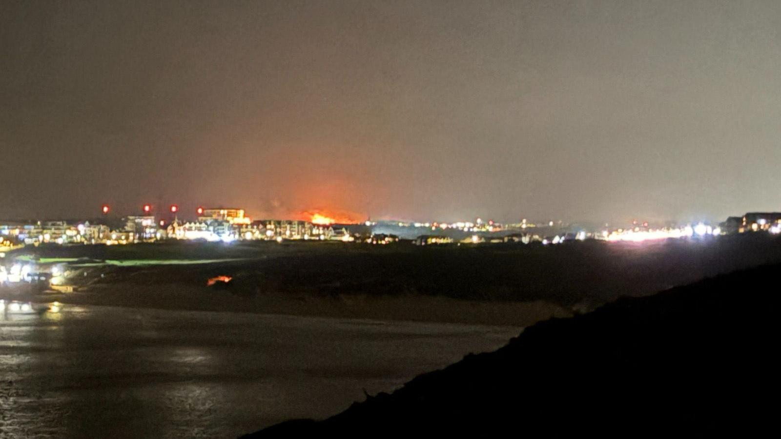 Flames from a fire on Rosenannon Downs seen in the distance from Newquay. A number of buildings with their lights on can be seen along Newquay's skyline. It is night time.