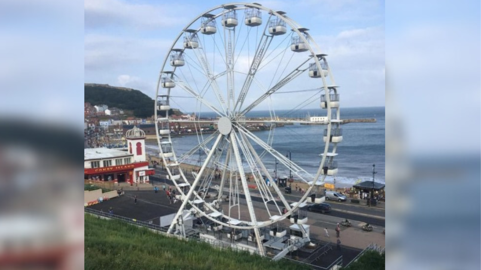 A large Ferris wheel occupies a site along Scarborough sea front. The wheel is white, with evenly spaced gondolas. Behind it, a view of the sea, the beach and the coastline can be seen, and to the left, a white building housing an amusement arcade.