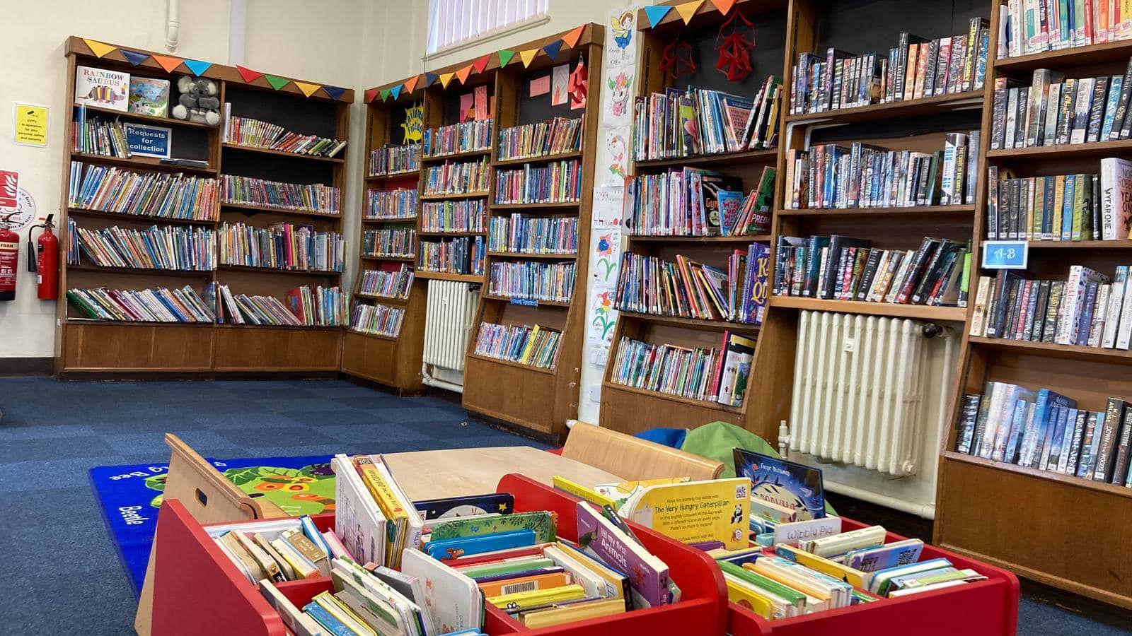 The interior of Woodston Library with rows of wooden shelves holding books. The room has a blue carpet and in the centre is a red box filled with children's books.  