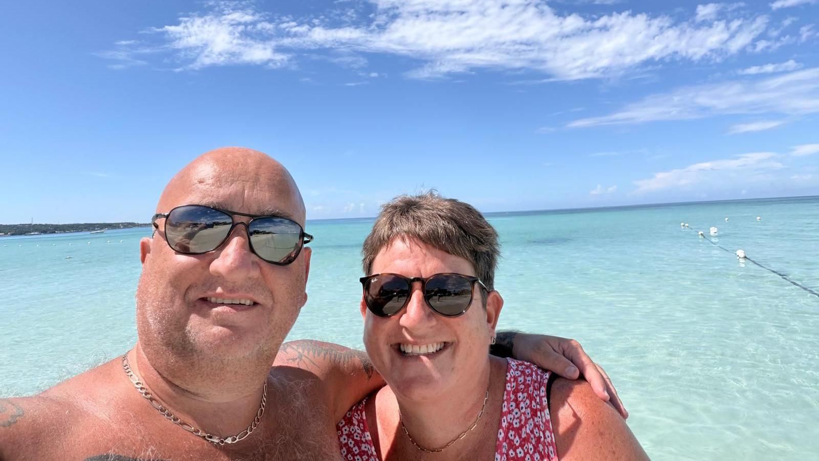 A couple are stood having a selfie in the ocean which looks blue and calm in the background. Its clear they are on some sort of island setting. The man is stood on the left with his arm around the women. They are both wearing sunglasses and smiling at the camera. 