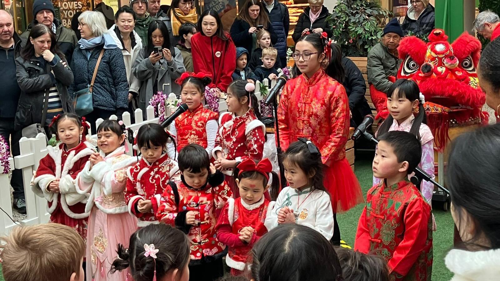 A group of children in red and pink outfits sing into microphones. A crowd watches around them.