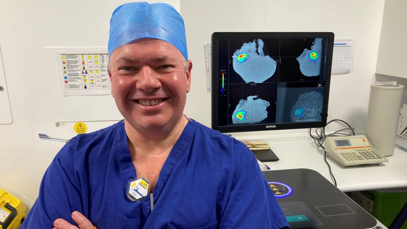 A man stands in front of a computer screen in a room with white walls. The man is wearing a blue surgical gown and a blue plastic cap that covers his hair. The computer screen behind him shows images from a medical scan, with green and red patches on a larger grey area.