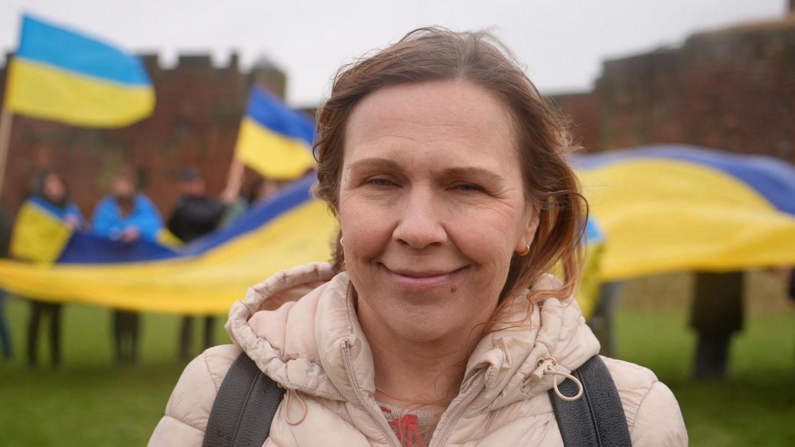 Olga Leintovea. She is smiling into the camera. She has brown hair and is wearing a pink jacket. Behind her are people at the march carrying blue-and-yellow Ukraine flags.