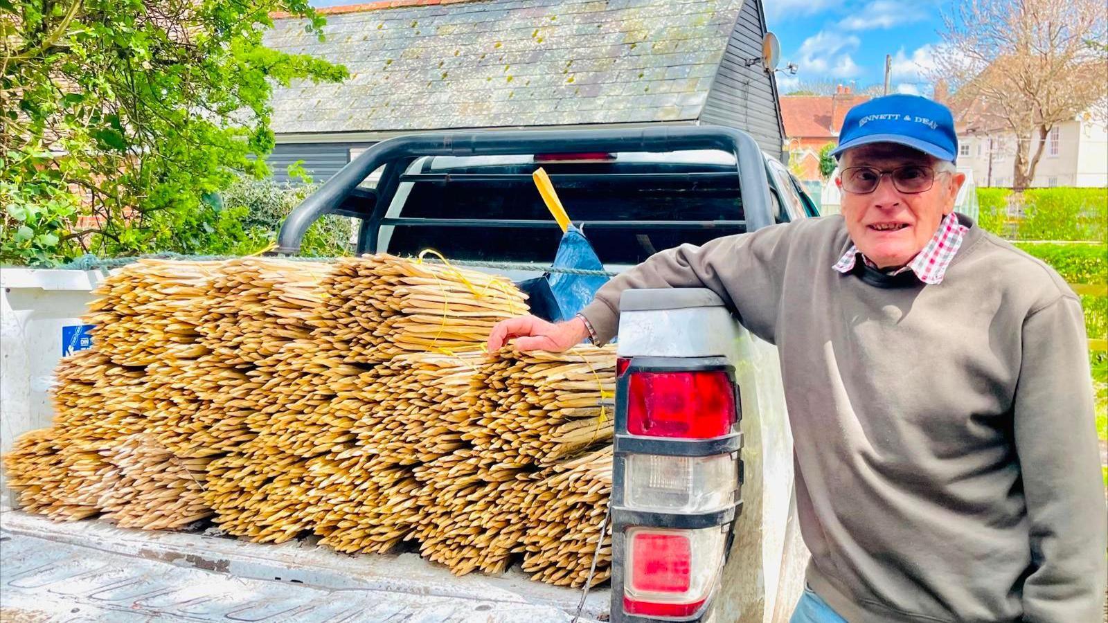 Ken Galton is standing at the back of a van with 14 bundles of sticks that will be used for British crafts. He is wearing a grey jumper and a blue cap. 