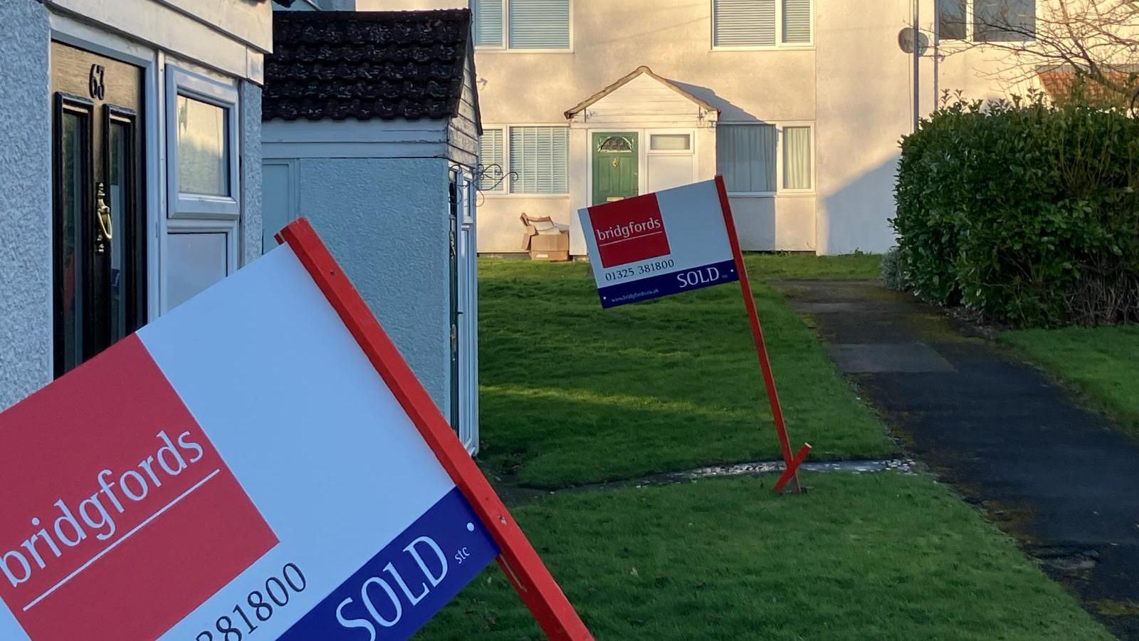 Two "sold" signs outside terraced houses in the foreground. Similar houses stand in the background, bathed in sunshine.