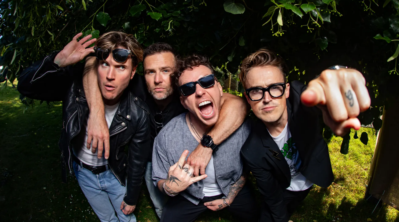 The four members of McFly in a close huddle in front of a tree, looking and point at the camera