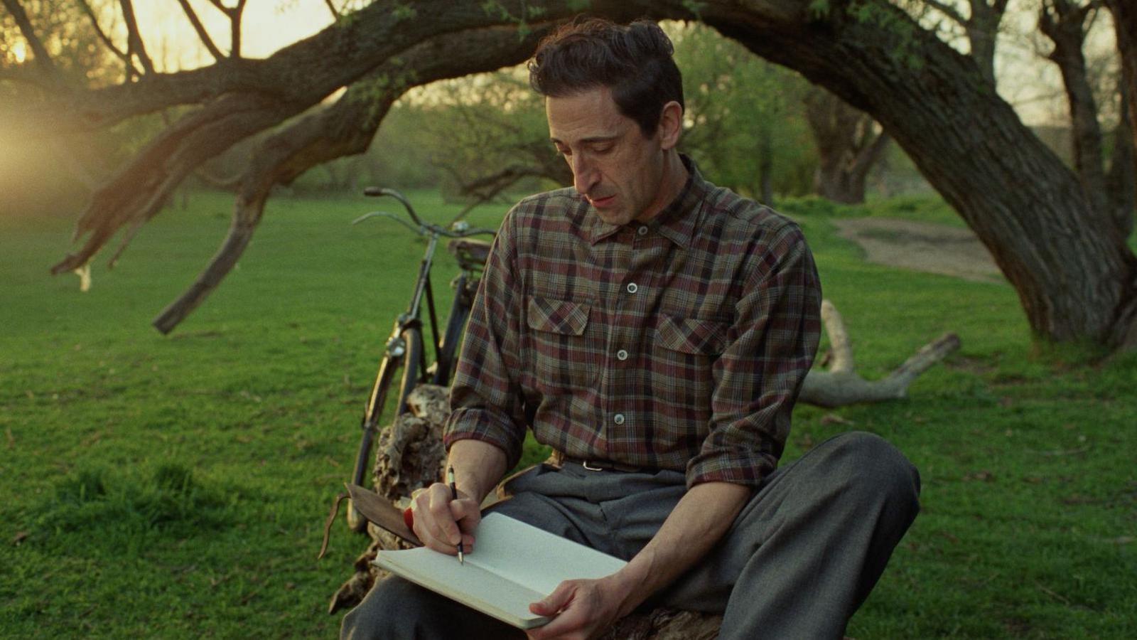 Adrien Brody, pictured as Tóth in a still from the film, is sitting under a tree, sketching. He is wearing a check shirt, buttoned to the neck, and grey trousers.