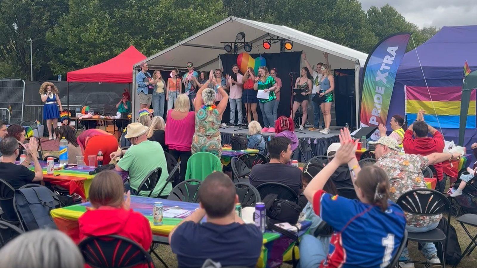 Thirteen people stand on the PrideXtra stage at Reading Pride. The stage is small and the people standing shoulder to shoulder fill its entire width. Scores of people are applauding while sitting and standing around tables decorated with rainbow tablecloths