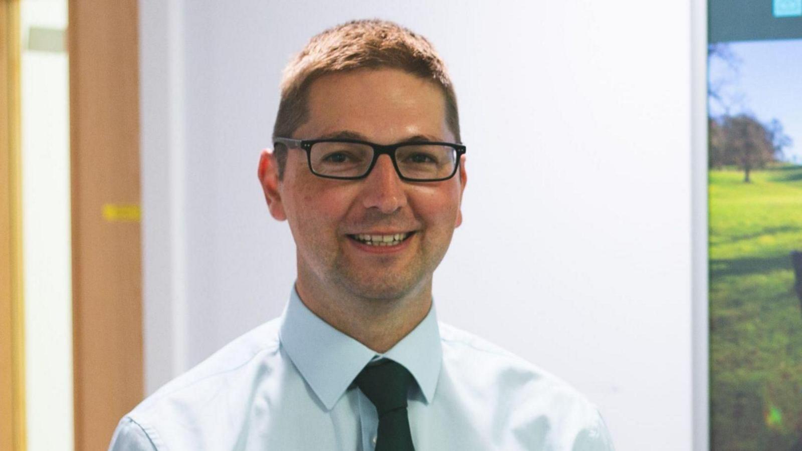 Councillor Richard Stanley wearing a white collared shirt, a black tie and black rectangular glasses. He has brown hair and is smiling at the camera, standing in front of a white wall in an office. 